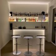 Angolo bar con banco illuminato, tre sgabelli e mensole retroilluminate con bottiglie
