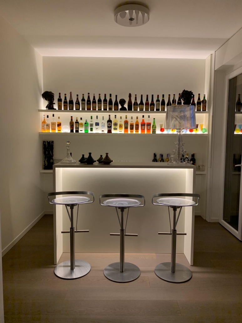 Attico Vigentino_01-PF-Archistudio Angolo bar con banco illuminato, tre sgabelli e mensole retroilluminate con bottiglie